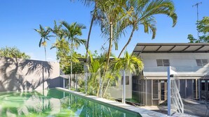 A swimming pool with clear water, surrounded by palm trees and a modern house with a covered patio.