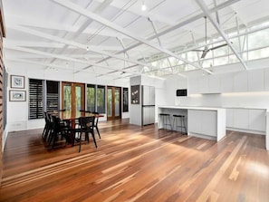 A spacious room with a wooden floor, a dining area with chairs, a kitchen with white cabinets, and a large window allowing natural light.