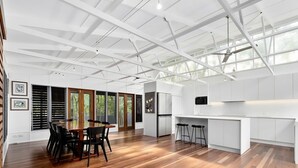 A spacious room with a wooden floor, a dining table with chairs, a kitchen area with white cabinets, and large windows allowing natural light.