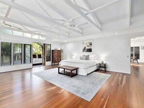 A spacious bedroom with a large bed, a wooden floor, a ceiling fan, and a view through the glass doors.