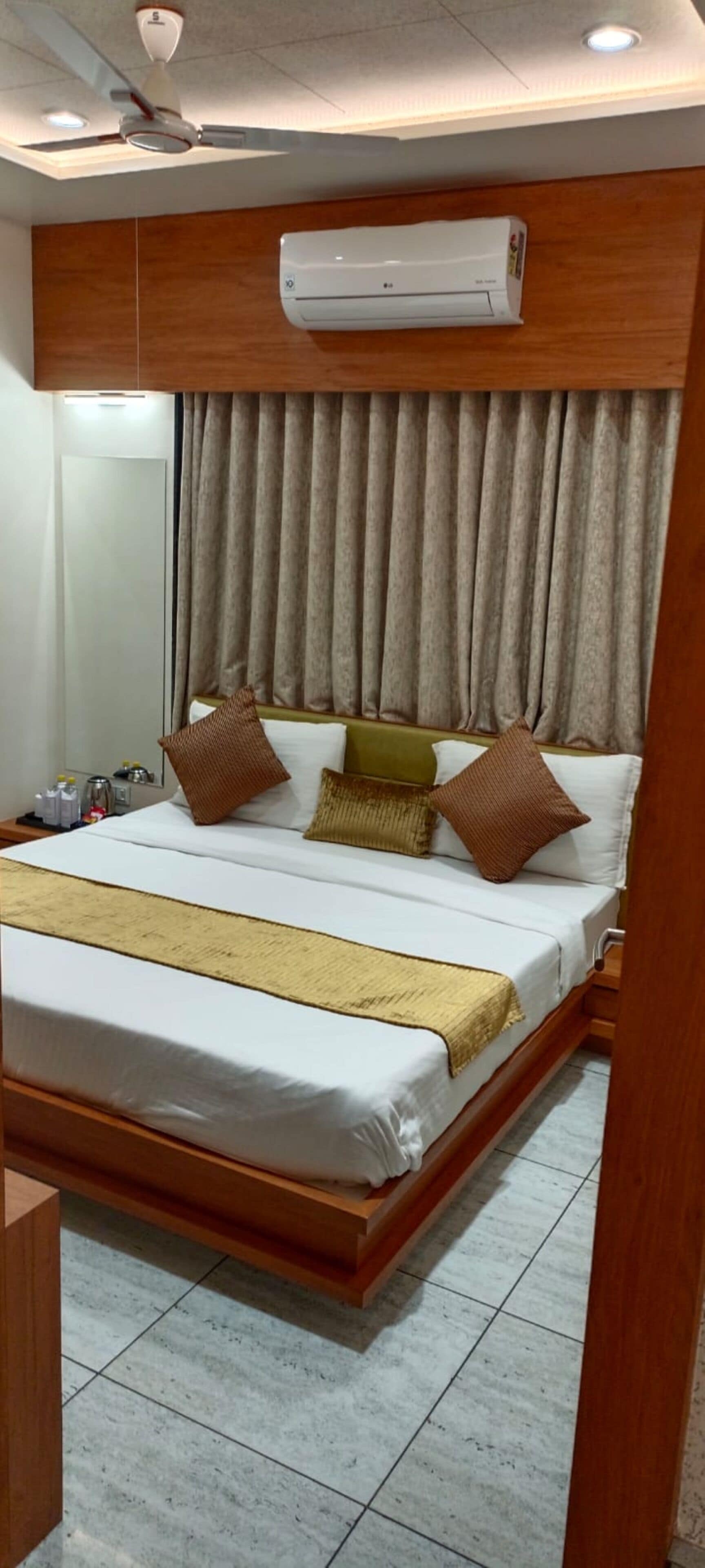 A hotel room with a large bed, wooden headboard, beige curtains, and an air conditioning unit above the bed.