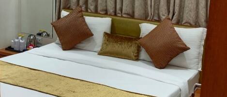 A hotel room with a large bed, wooden headboard, beige curtains, and an air conditioning unit above the bed.