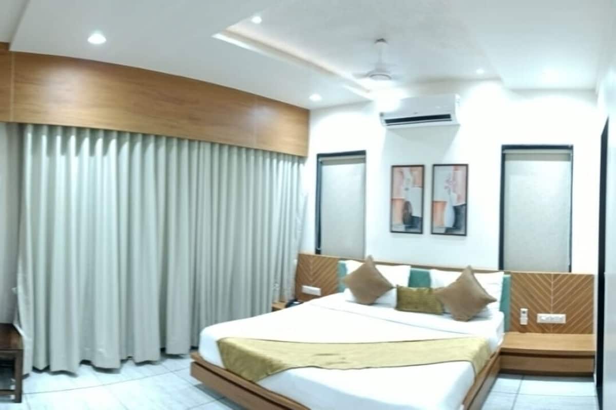 A modern hotel room with a large bed, a wooden headboard, a small bedside table with toiletries, and a large window with curtains.
