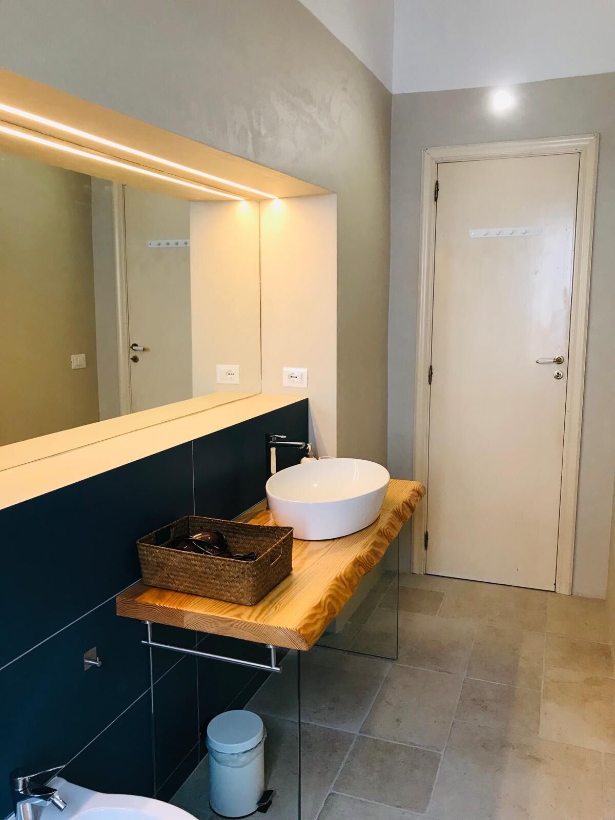 A modern bathroom with a large mirror, a wooden vanity, a white sink, and a small trash can.
