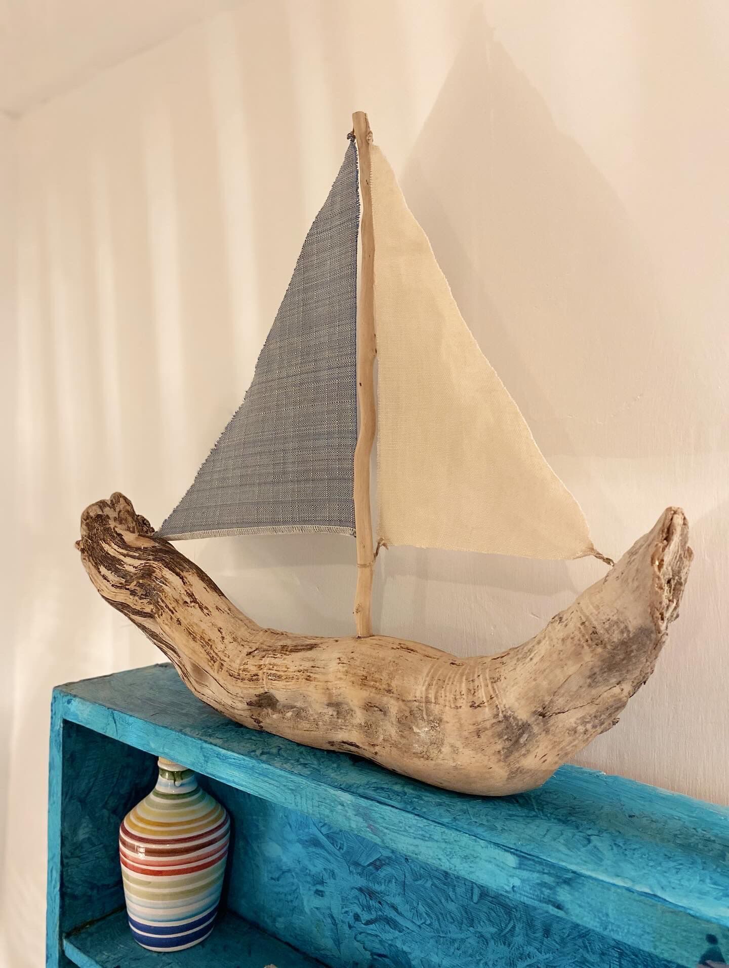 Un velero modelo sobre un pedazo de madera a la deriva, sobre un estante azul con un florero a rayas debajo.
