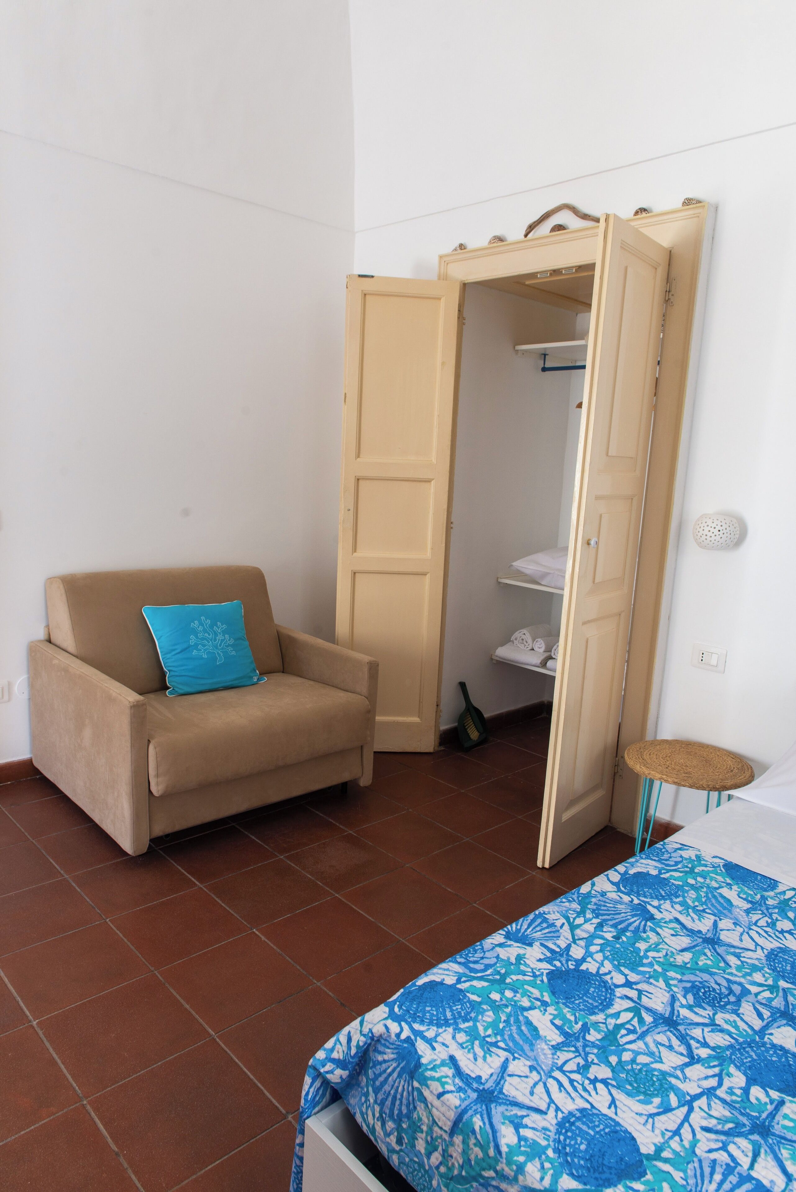 A room with a beige armchair, a blue patterned bedspread, a wardrobe, and a small stool.
