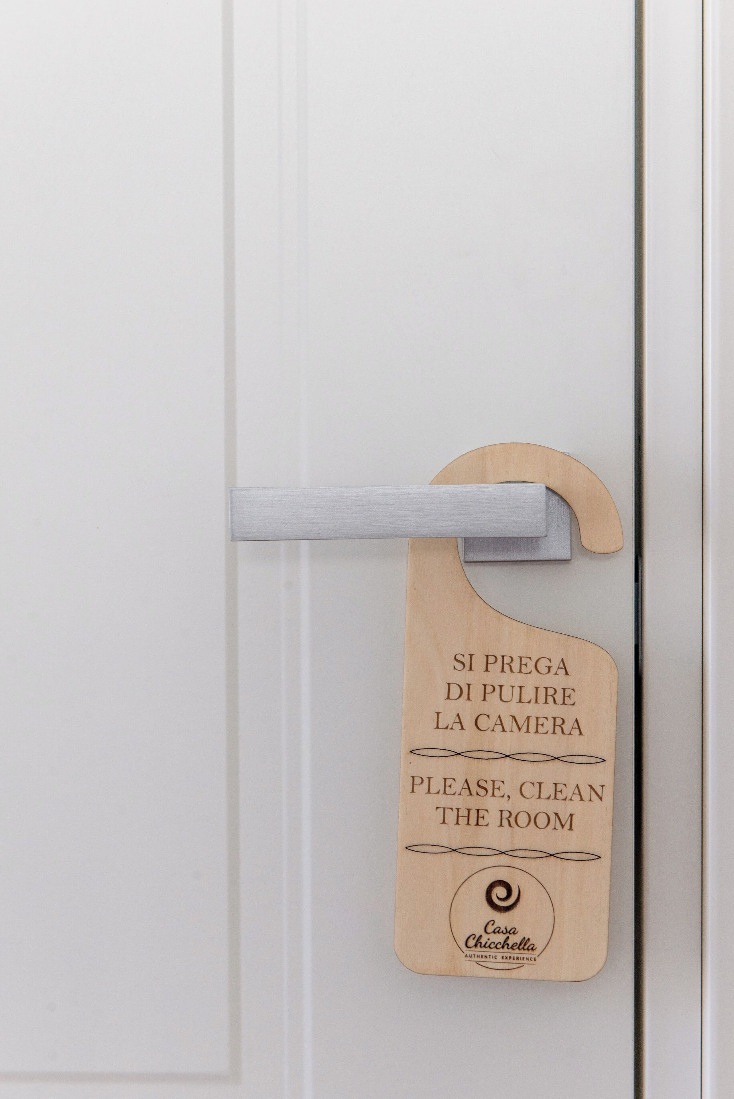 Ein hölzernes Schild an einer Tür mit Text auf Italienisch und Englisch, das darauf hinweist, den Raum zu reinigen, falls die Türklingel gedrückt wird.