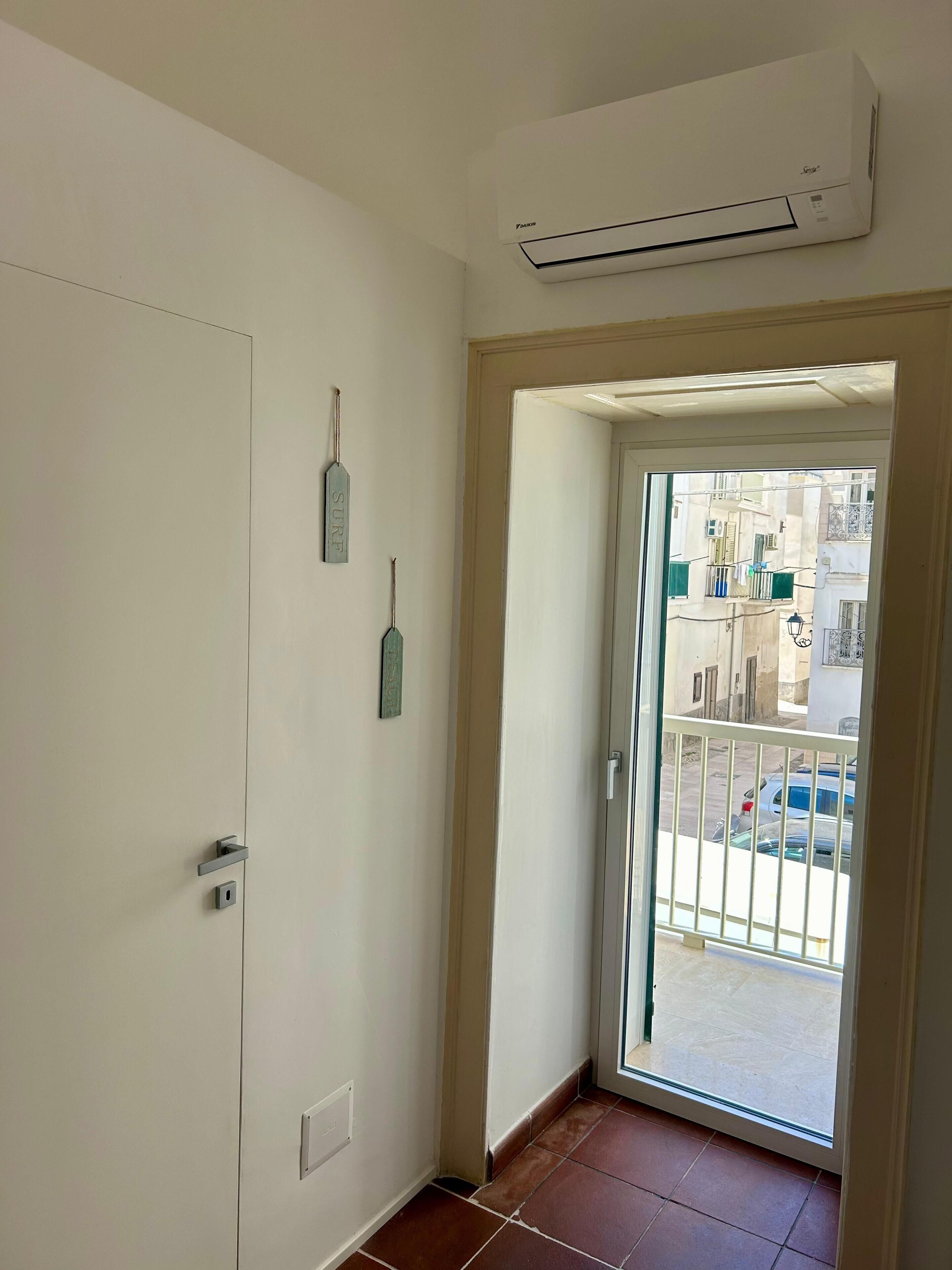 A narrow hallway with a white door, a small window, and a view of a balcony with a railing and parked cars.