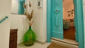 A hallway with a teal door, a green vase with pampas grass, a wicker basket, and a small white shelf with a picture frame. - Casa Chicchella (Vieste)