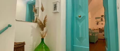 A hallway with a teal door, a green vase with pampas grass, a wicker basket, and a small white shelf with a picture frame.