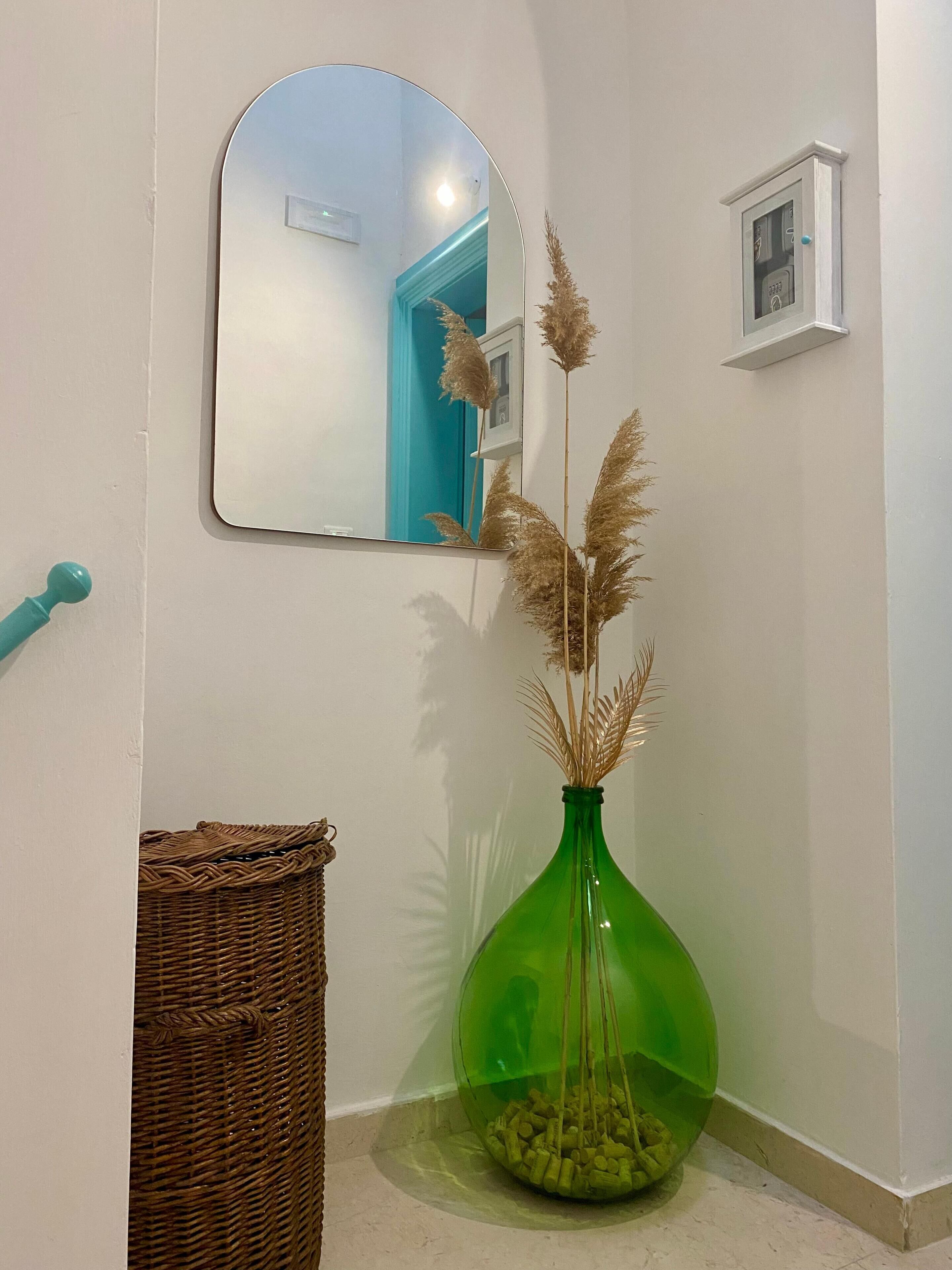 A hallway with a green vase containing pampas grass, a wicker basket, and a mirror reflecting a blue door.