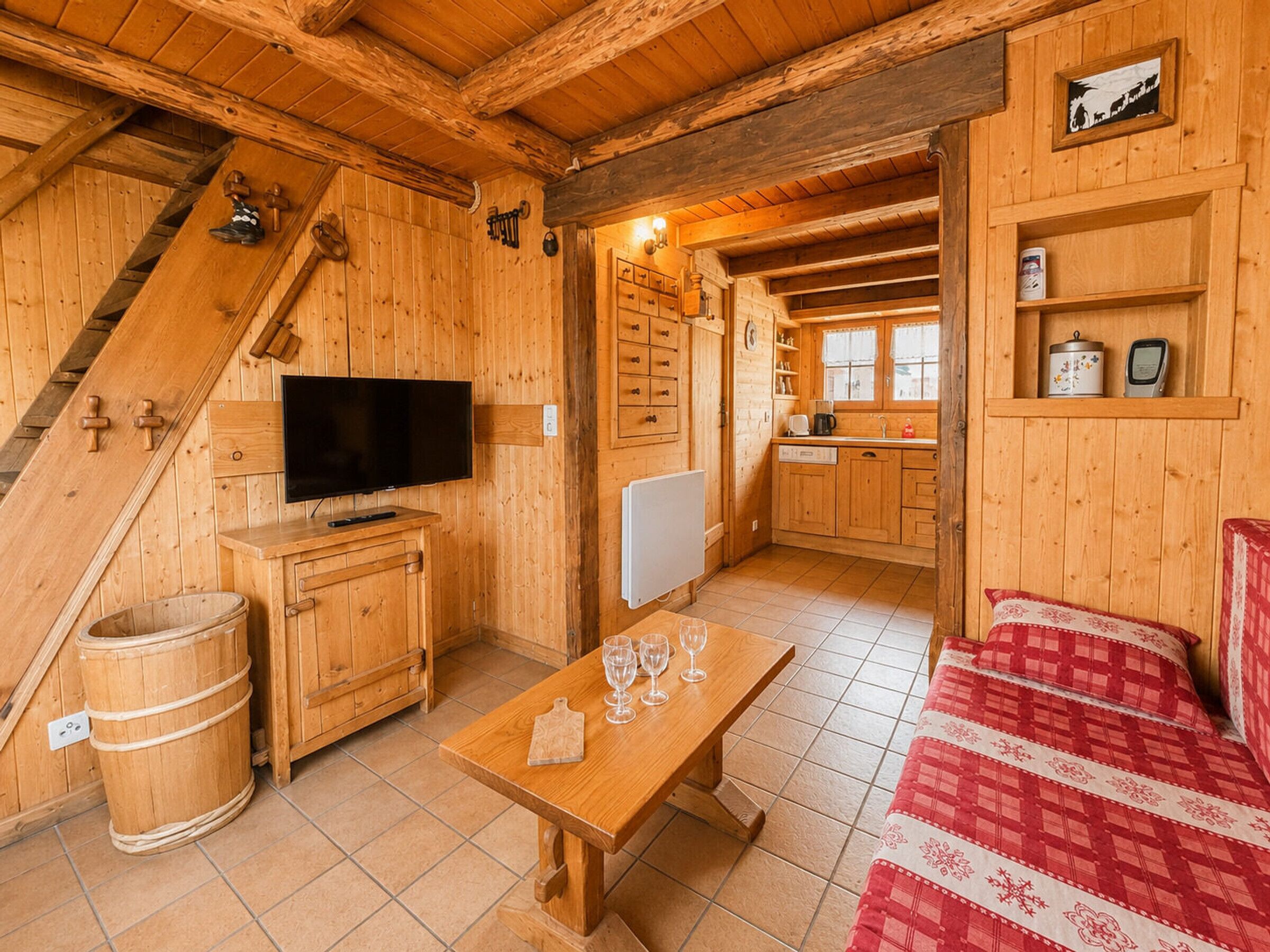 Un intérieur en bois avec un téléviseur, des meubles en bois, une table à manger avec des verres, un canapé rouge à carreaux et un coin cuisine.