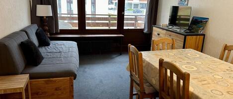 Una habitación compacta con sofá, una pequeña mesa de madera, una mesa de comedor con sillas, televisión y ventana con vista a edificios.