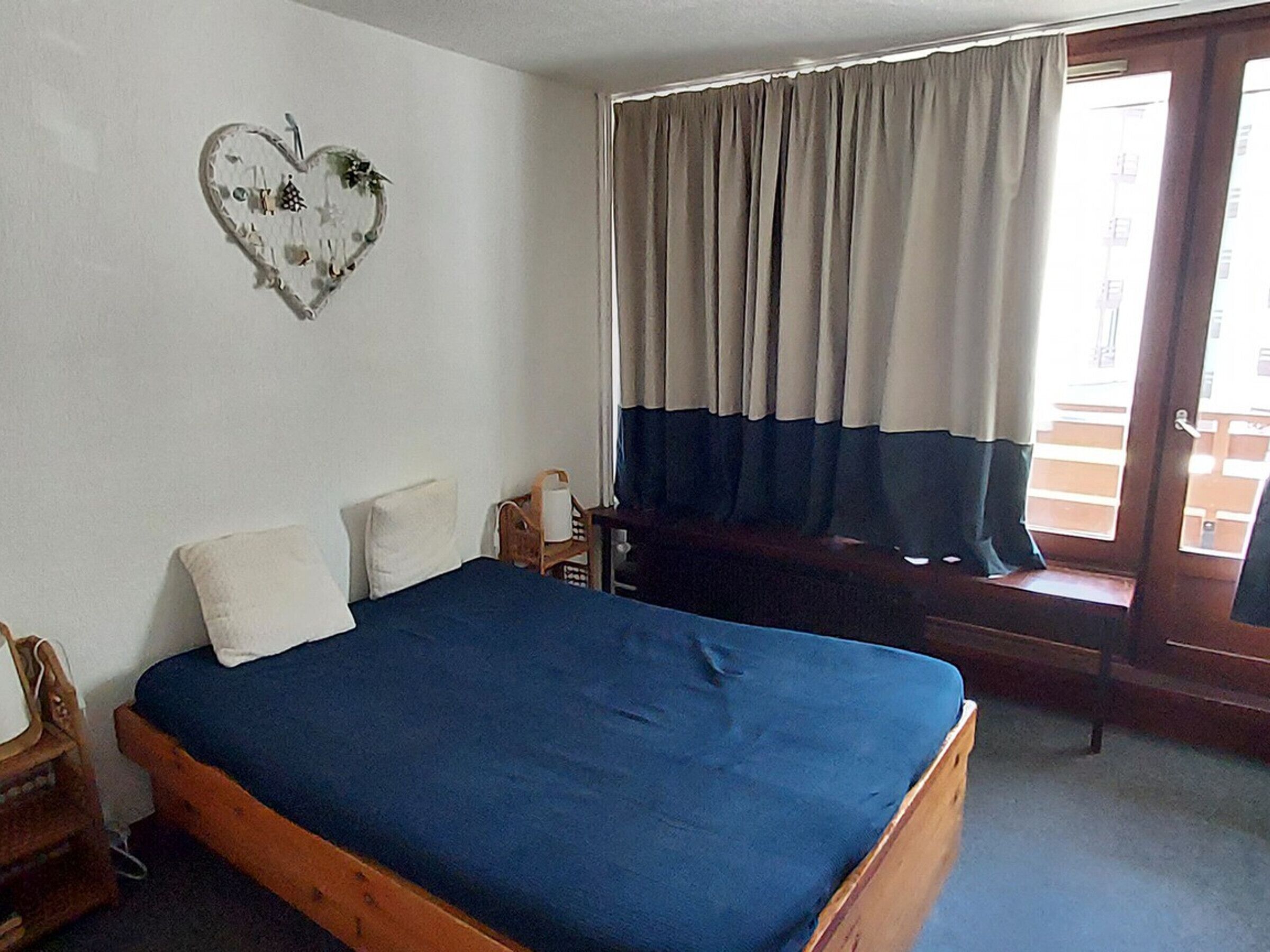 A bedroom with a wooden bed, a white wall, a heart-shaped decoration, and a window with curtains.