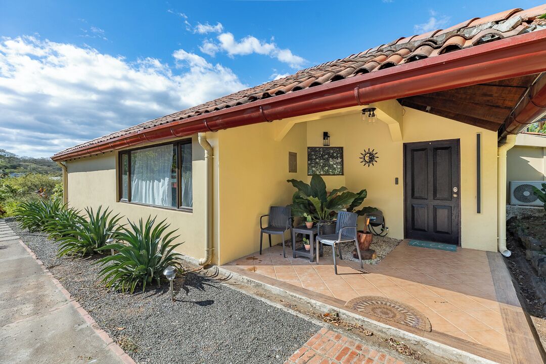 A single-story house with a tiled roof, a front porch with chairs, and a door.
