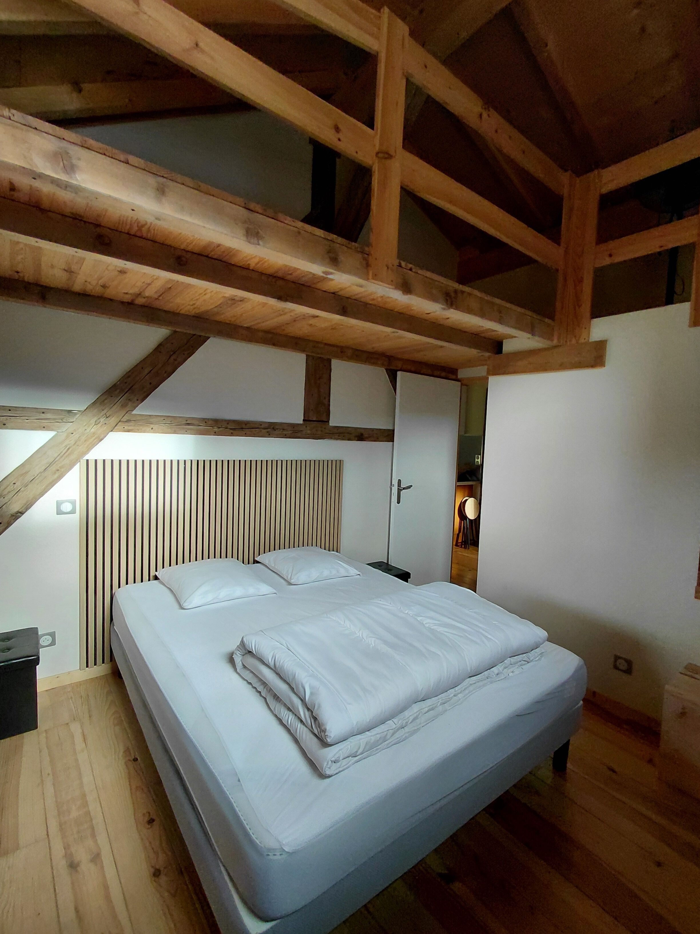 Une chambre avec un plafond en bois, un lit avec une literie blanche, une table de chevet et une lampe fixée au mur.