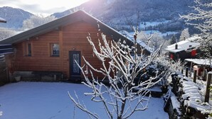 A wooden cabin with a snow-covered yard and mountains in the background.