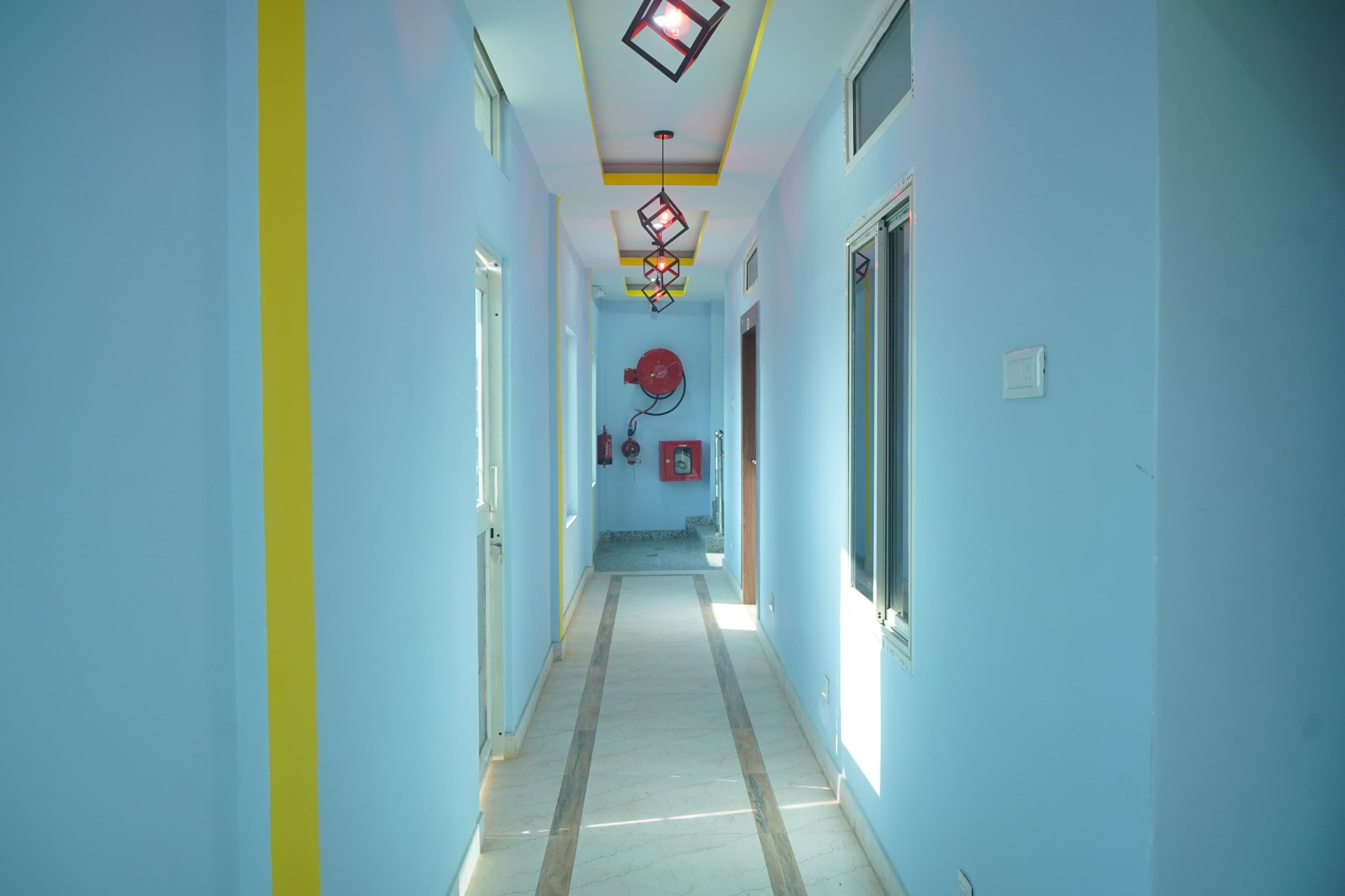 Un pasillo estrecho con paredes azul claro, una franja amarilla y suelo de baldosas. Hay un extintor y un ventilador rojo en la pared.