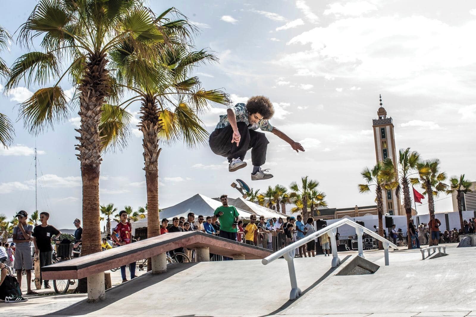 A skateboarder performing a trick at a skate park with palm trees and a tower in the background.