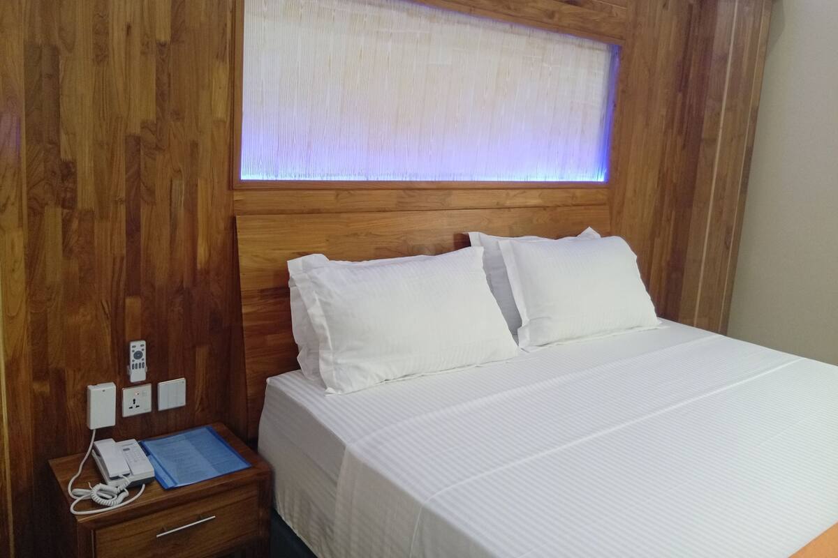Een houten hoofdbord met ingebouwde verlichting, een wit bed met twee kussens, een nachtkastje met een telefoon en een blauwe map, en een stopcontact.