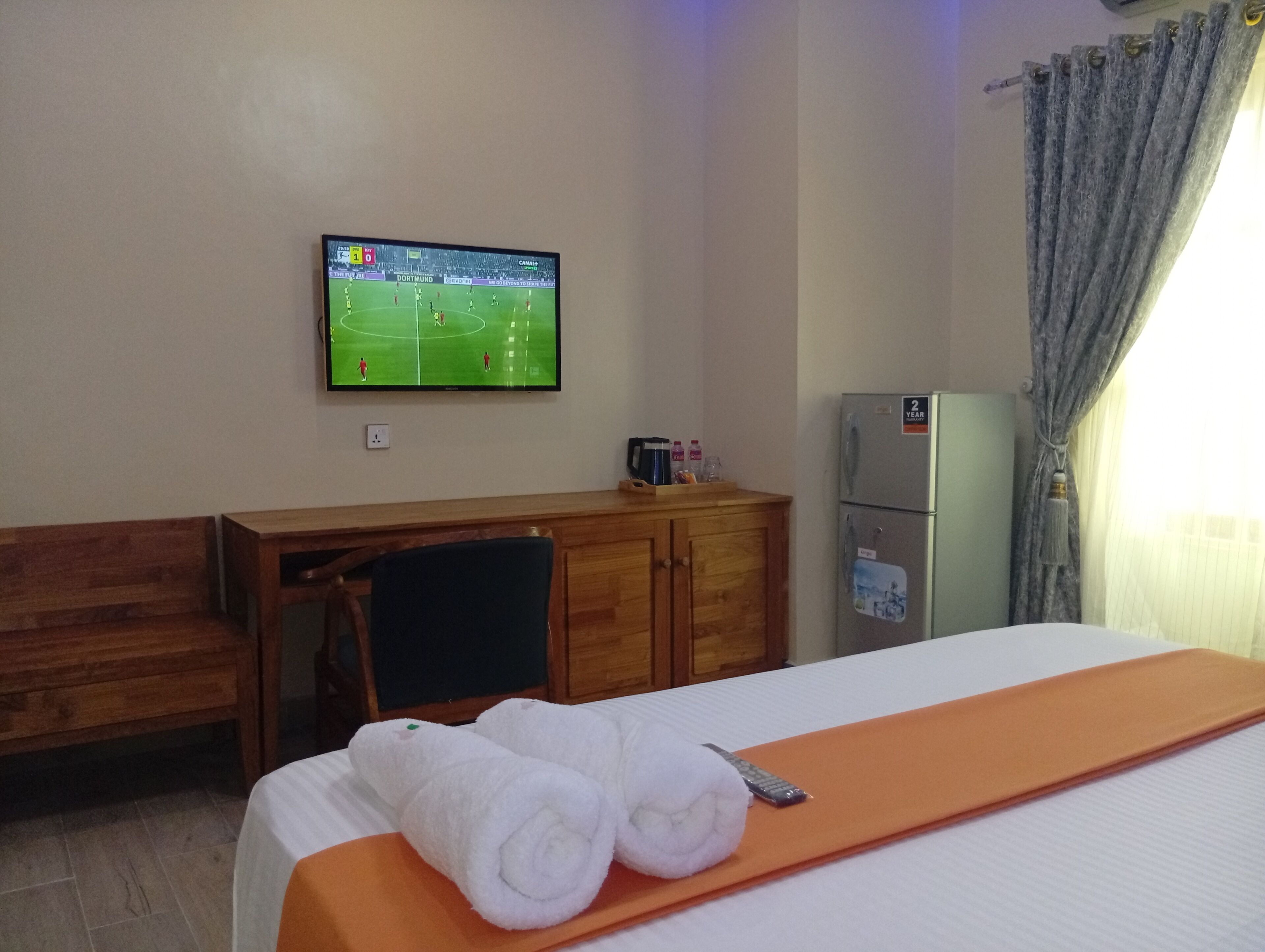 A hotel room with a bed, a desk, a television displaying a soccer match, a refrigerator, and a window with curtains.