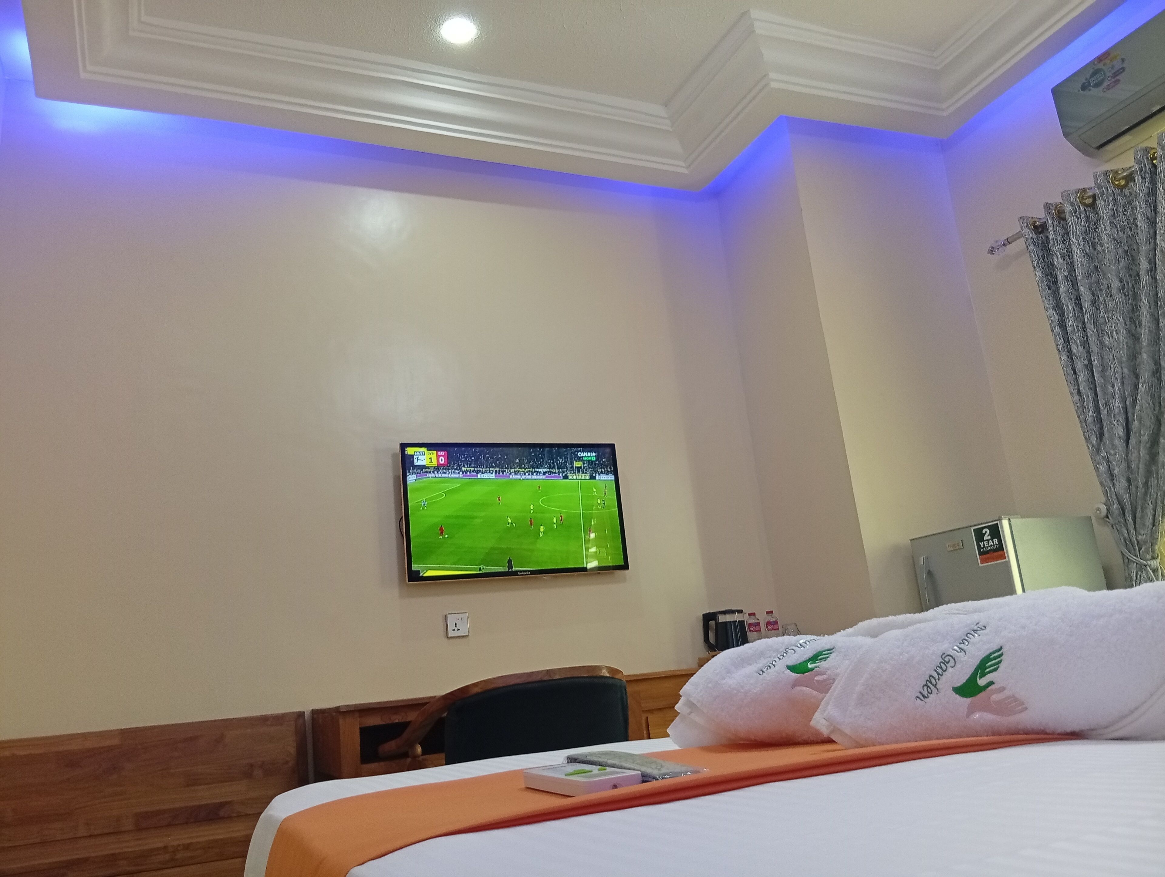 Une chambre d’hôtel avec un lit, un téléviseur diffusant un match de football, un petit radiateur et une fenêtre avec des rideaux.