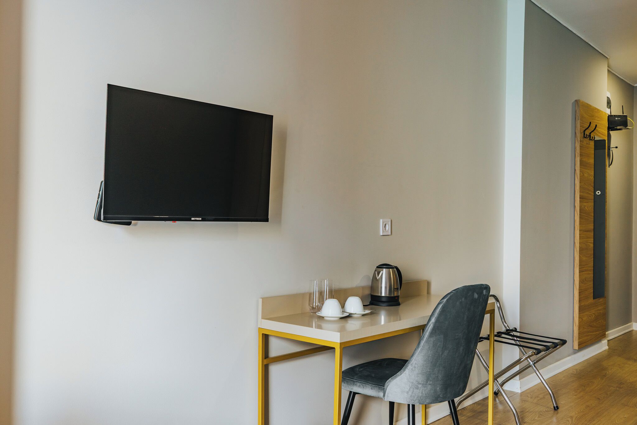 Une chambre moderne dotée d’un téléviseur à écran plat fixé au mur, d’une petite table en bois avec une bouilloire, des tasses et des verres, et d’une chaise grise.