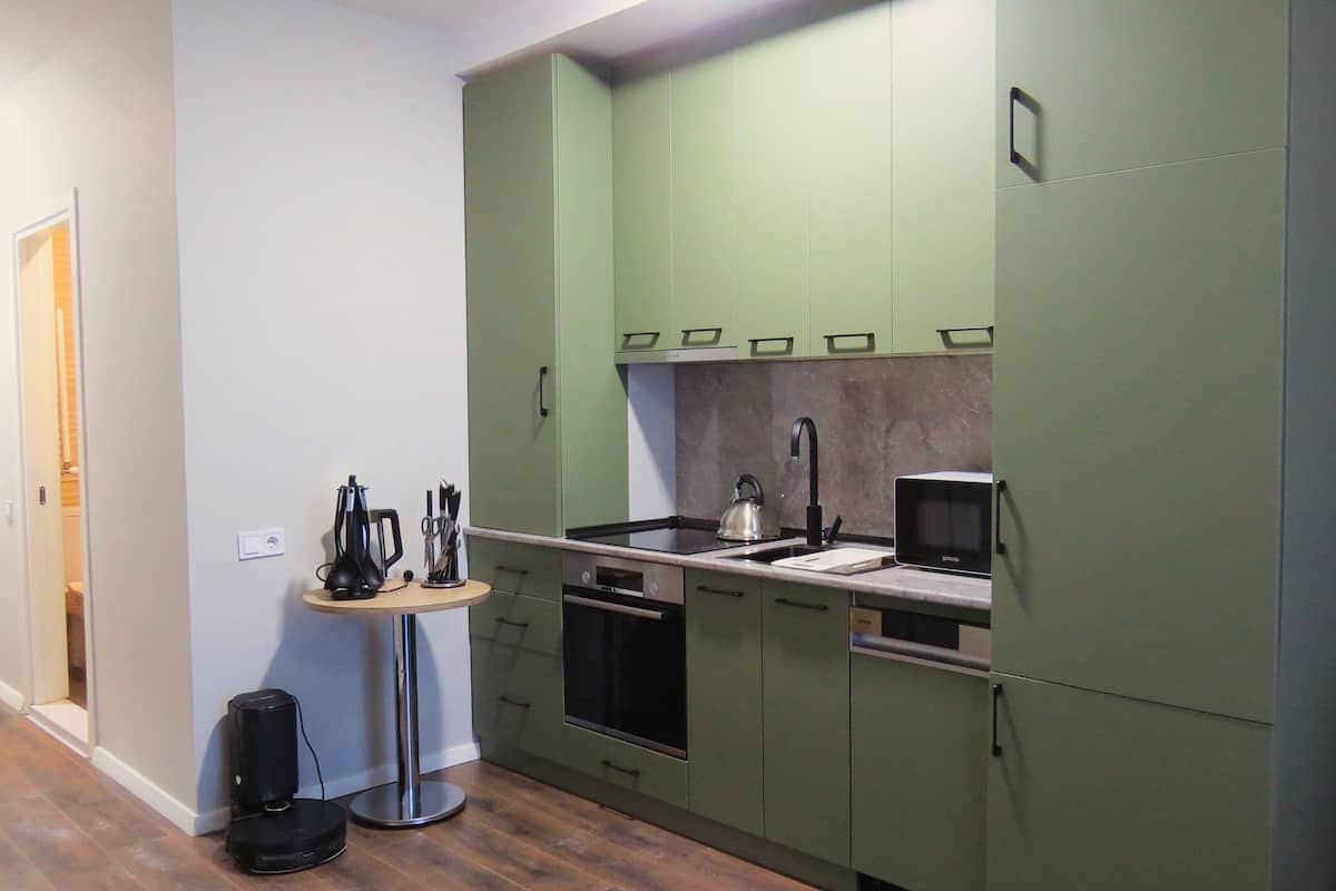 A compact kitchen with green cabinets, a microwave, a kettle, and a small round table with a speaker and utensils.