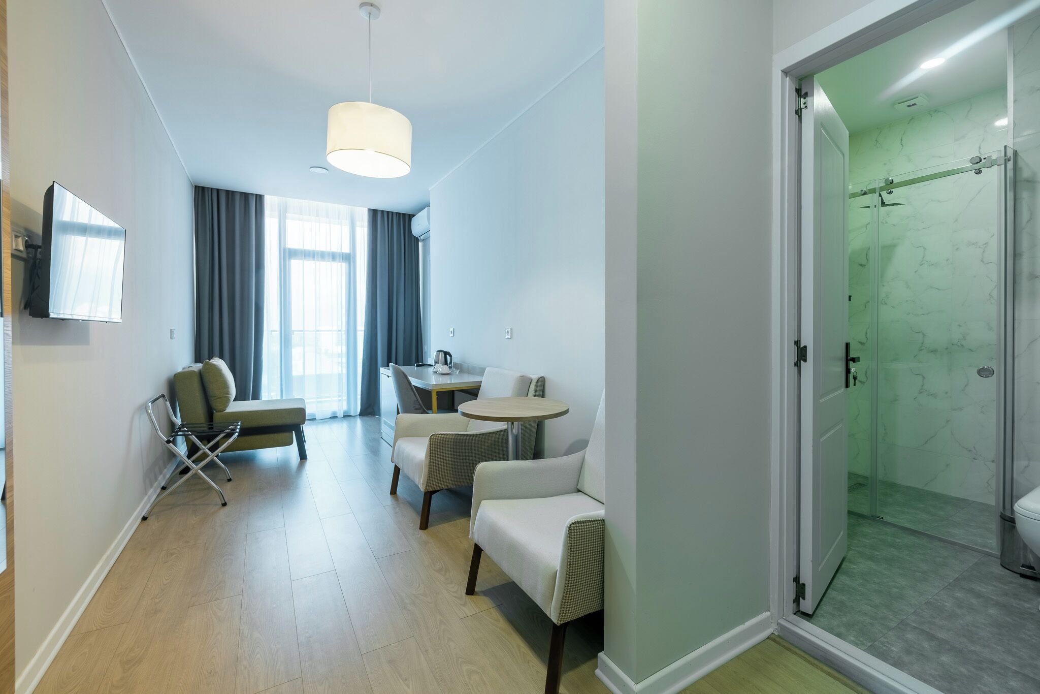 A modern hotel room with a seating area, a dining table, a shower with glass doors, and a television mounted on the wall.