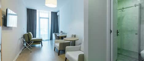 A modern hotel room with a seating area, a dining table, a shower with glass doors, and a television mounted on the wall.