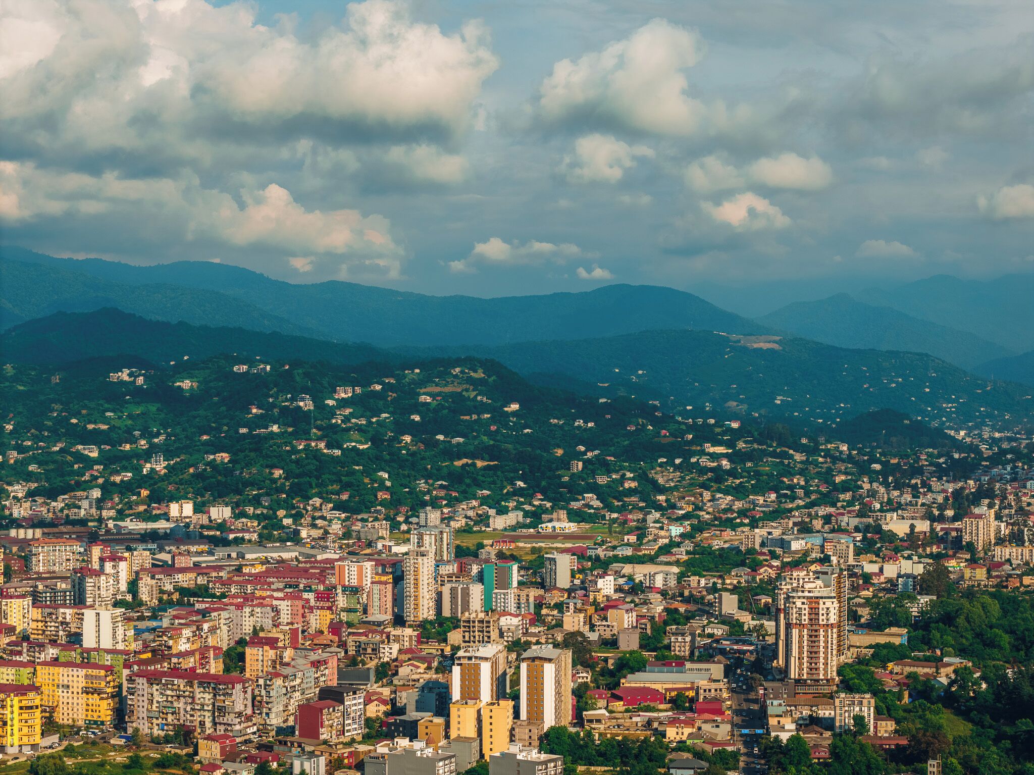 Un paisaje urbano con edificios densos, montañas al fondo y zonas verdes dispersas.