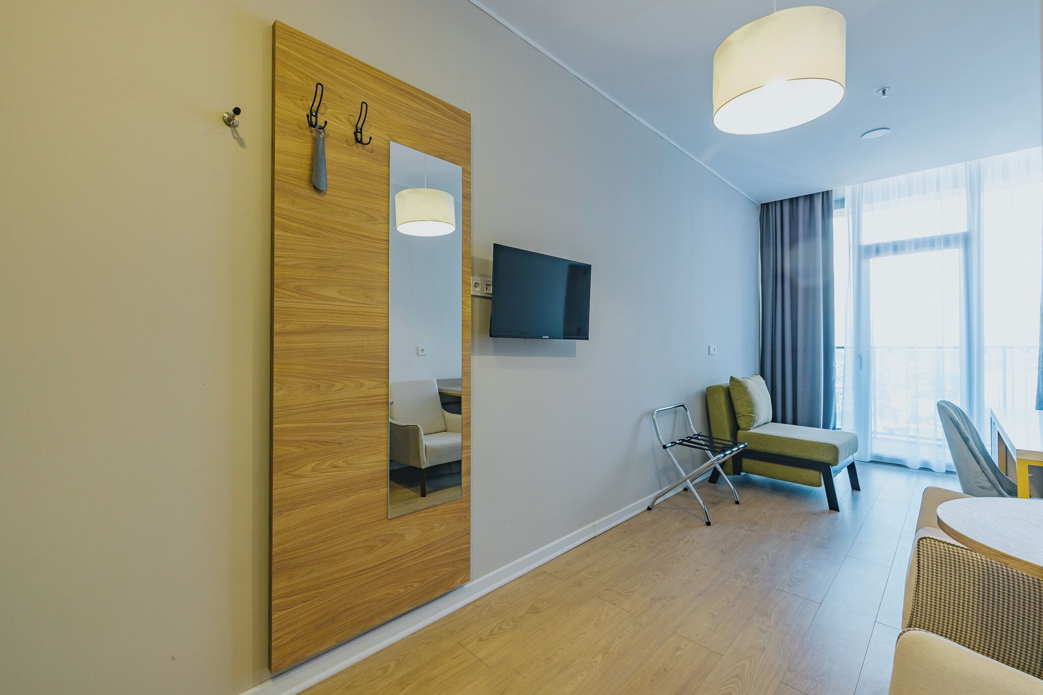 Et moderne hotelværelse med et træskab, et fladskærms-tv, en sofa, et spisebord og et stort vindue med gardiner.