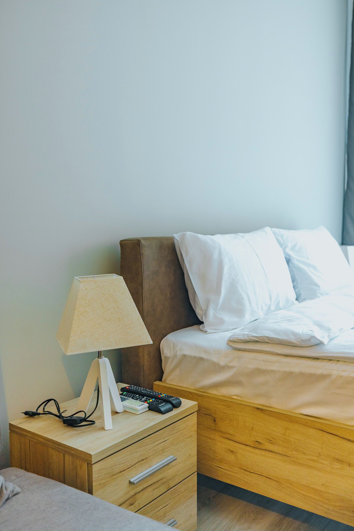 A neatly made bed with white bedding, a wooden nightstand with a lamp, a remote control, and a phone charger.