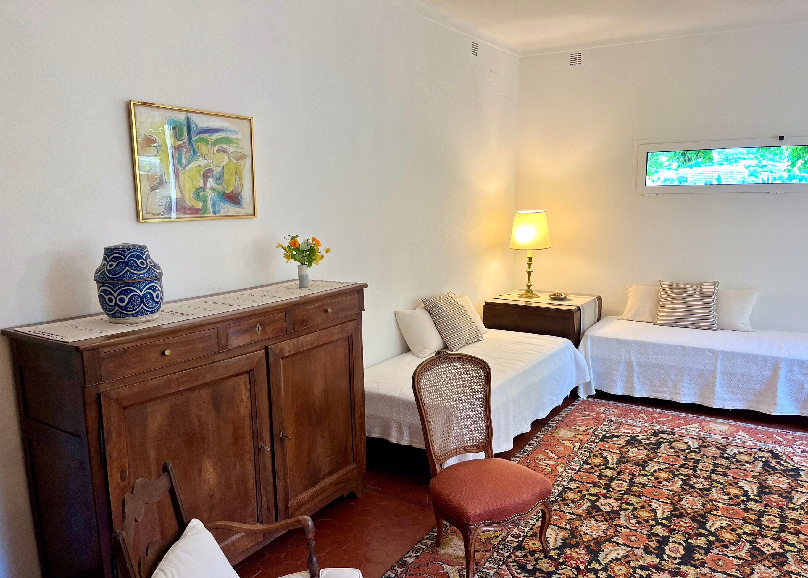 Quarto de hotel com duas camas, uma cômoda de madeira, uma cadeira, um abajur, um vaso com flores e um quadro na parede.