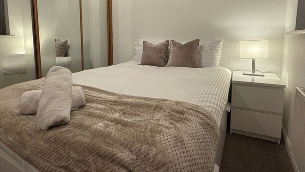 A neatly made bed with a beige blanket and pillows, a bedside table with a lamp, and a mirror on the wall.