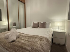 A neatly made bed with a beige blanket and pillows, a bedside table with a lamp, and a mirror on the wall.