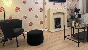 A living room with a fireplace, a black armchair, a round ottoman, a glass coffee table, and a flat-screen TV displaying 'NETFLIX'.
