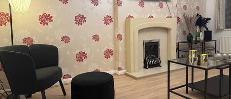 Una sala de estar con chimenea, un sillón negro, un reposapiés redondo, una mesa de centro de vidrio y un televisor de pantalla plana mostrando 'NETFLIX'.