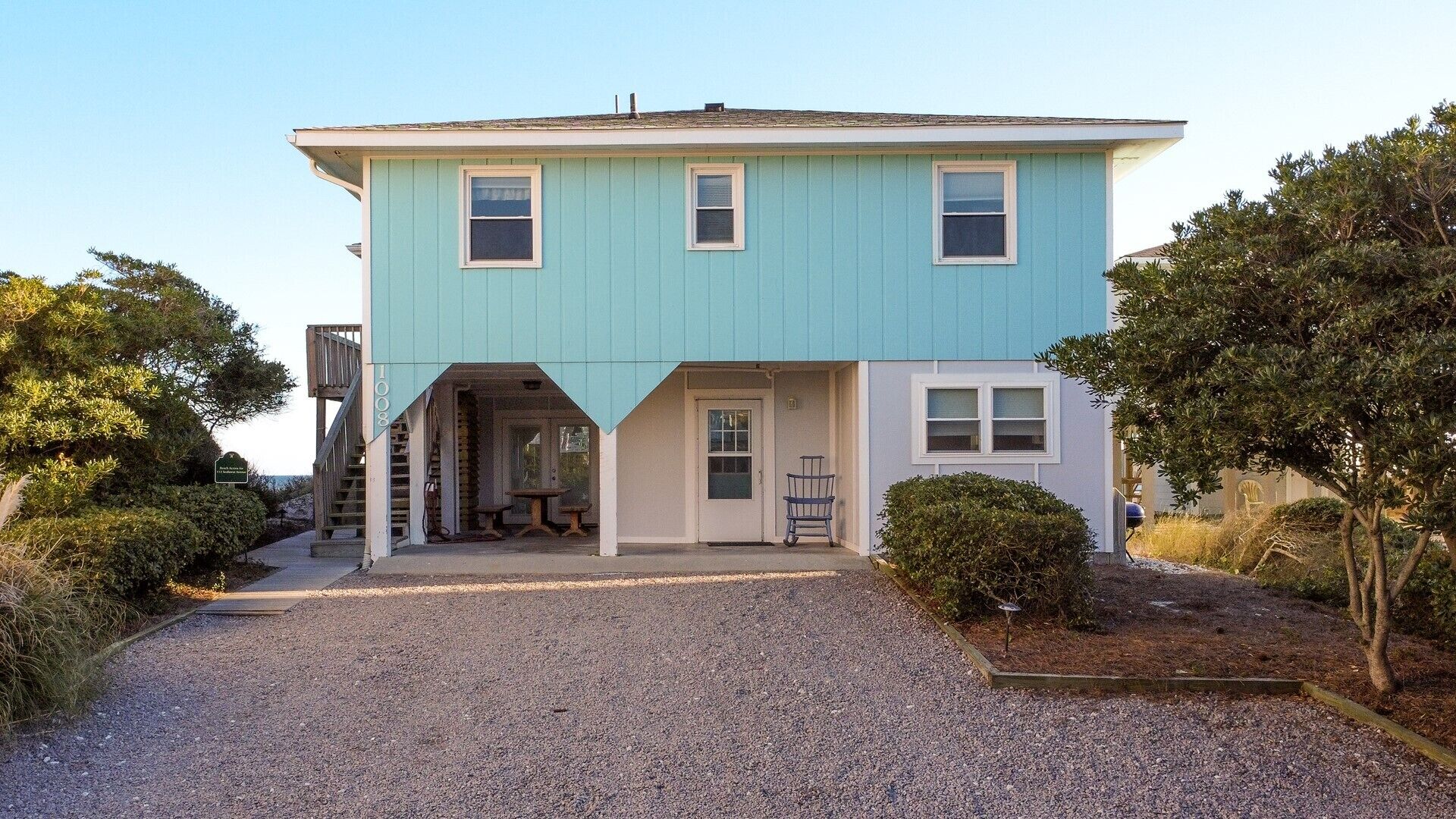 A two-story house with a porch, a staircase leading to a deck, and a gravel driveway.