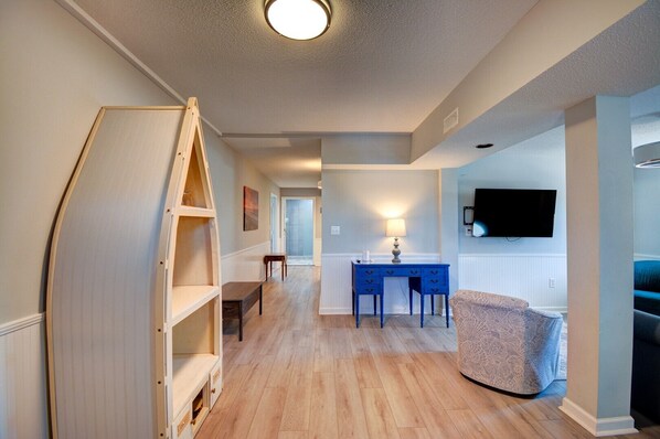 A hallway with a wooden ladder, a desk with a lamp, a television mounted on the wall, and a patterned sofa.