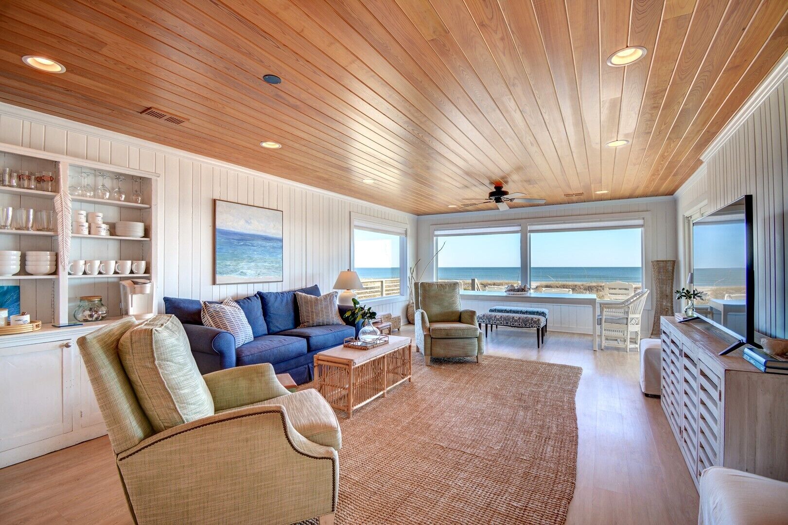 Een ruime woonkamer met een groot raam met uitzicht op het strand, een gezellige zithoek en een houten plafond.