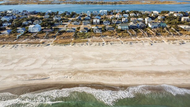 Vue aérienne d’un quartier résidentiel situé sur la côte, avec des maisons alignées le long de la plage et des vagues qui se rapprochent du rivage.