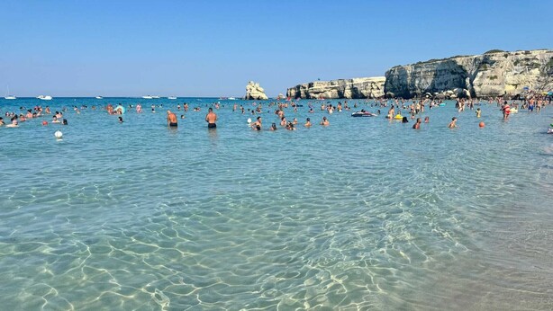 A beach with clear shallow waters, rocky cliffs, and a crowd of people.
