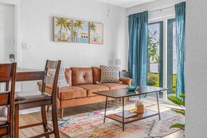 Un salon avec un canapé en cuir marron, une table basse en bois, un tapis coloré et une vue sur l’extérieur grâce à de grandes fenêtres ornées de rideaux bleus.