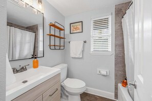 A bathroom with a white sink, toilet, and shower. There is a framed picture on the wall and a window with blinds.
