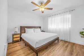 A bedroom with a wooden bed, a nightstand, a lamp, a ceiling fan, and a window with sheer curtains.