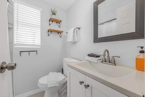 Une salle de bain équipée d’un WC, d’un lavabo, d’un miroir, d’un porte-serviettes et d’une fenêtre avec des stores.