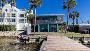 Een kusthuis met een houten steiger naar een turquoise woning met een balkon, omringd door palmbomen en een helderblauwe lucht.