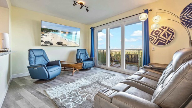 A living room with a sofa, two armchairs, a coffee table, a patterned rug, a painting of a beach, and a balcony with a view. - RIVER FLY IN: Updated, Luxurious Waterfront 2BR (Merritt Island)