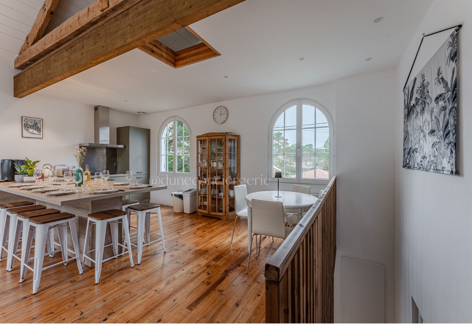 A spacious kitchen with wooden beams, a dining area with white chairs, and large windows offering views of greenery.